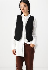 Svart skreddersydd vest med en tettsittende design, seks knapper og lommer, brukt over en hvit skjorte med knapper og brune kordfløyelsbukser.
