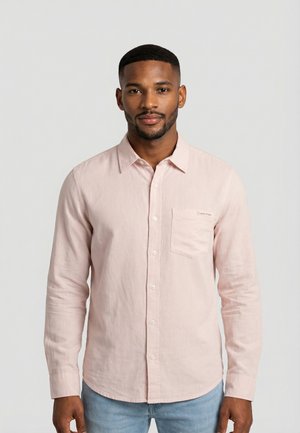 Uomo con camicia rosa chiaro a bottoni e tasca frontale, e jeans azzurri, in piedi davanti a uno sfondo grigio neutro.