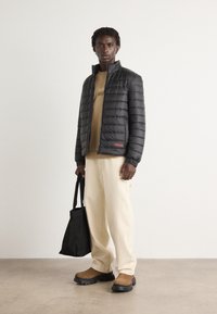 Zwarte pufferjack met quilted textuur, ritssluiting en rood merklogo; beige T-shirt eronder, crèmekleurige broek en bruine schoenen.