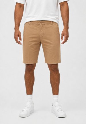 Hombre con pantalones cortos caqui hasta la rodilla, calcetines blancos, zapatillas blancas y una camiseta blanca metida por dentro, de pie frente a un fondo liso.