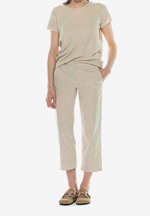 Kobieta ubrana w beżowe spodnie o długości do kostek, brązowy t-shirt z krótkim rękawem i brązowe mokasyny wsuwane z złotym łańcuszkiem, trzyma rękę w kieszeni.