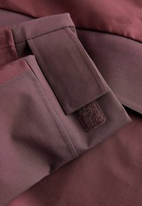 Nærbillede af maroon stof med en syet klaffem og et rektangulært velcro-patch på et beklædningsstykke.