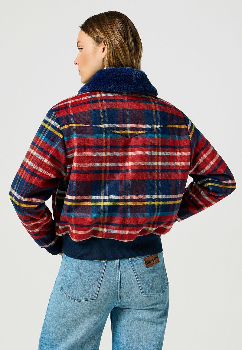 Giacca a quadri rossi e blu con colletto in pelliccia sintetica blu navy, orlo e polsini a coste. Presenta dettagli di cucitura sottili e un piccolo logo sulla tasca posteriore.