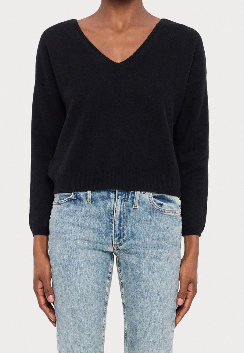 Woman wearing a black V-neck long sleeve sweater paired with light blue high-waisted jeans against a white background.