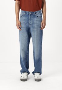 Personne portant un jean droit bleu clair avec un léger effet délavé et des baskets blanches avec des détails gris, debout sur un fond blanc.