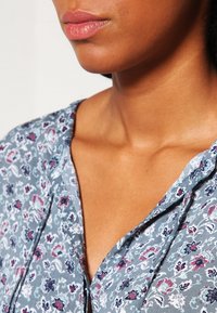 Blouse à motif floral en gris clair avec des accents roses et bleus. Tissu doux, col en V, coupe décontractée avec des boutons à l'avant.