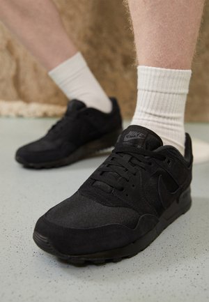Osoba nosząca białe skarpetki i czarne buty sportowe Nike stojąca na jasnej, cętkowanej podłodze na rozmytym brązowym tle.