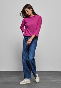 Sudadera magenta con un patrón texturizado, mangas tres cuartos, combinada con vaqueros de talle alto azules y zapatillas beige.