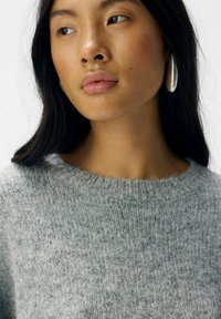 Hellgrauer gestrickter Pullover mit rundem Halsausschnitt und einem strukturierten Muster. Das Model trägt eine große silberne Creole. Glatte Hautfarbe.