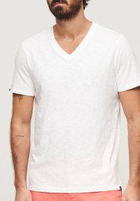 Biała koszulka z dekoltem w serek, wykonana z lekkiego materiału. Posiada luźny krój i krótkie rękawy, subtelną teksturę oraz małe logo z boku.