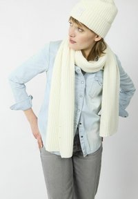 Bonnet tricoté blanc avec un pompon et un patch, associé à une longue écharpe à côtes assortie, porté sur une chemise en chambray bleu clair.