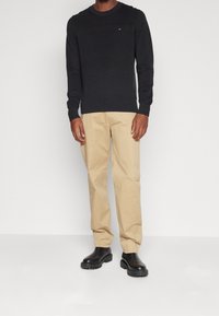 Schwarzer strukturierter Pullover, beige Twill-Hosen und schwarze Stiefel mit klobiger Sohle. Einfaches Design mit sichtbarem Logo der Marke auf dem Pullover.