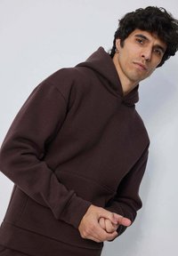 Hoodie marron en mélange de coton, avec une poche frontale, des poignets côtelés et une capuche spacieuse. Texture lisse sans motifs visibles.
