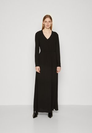 Black long-sleeve dress with a V-neck and gathered waist, made from a smooth fabric. Worn with pointed black ankle boots.