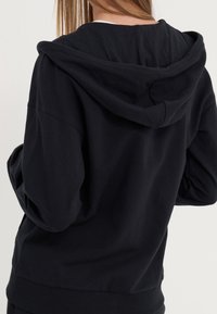 Sweat-shirt à capuche noir avec manches longues, ourlet côtelé et texture douce. La capuche est posée à plat sur le dos, mettant en valeur un design simple.