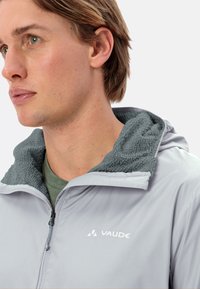 Veste gris clair avec une capuche en polaire texturée gris foncé, fermeture éclair à l'avant et logo VAUDE blanc sur la poitrine. Tissu lisse et coupe décontractée.
