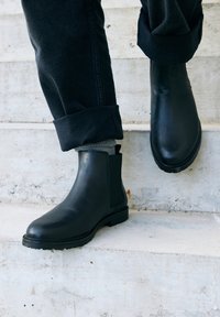 Schwarze Leder-Chelsea-Boots mit seitlichen elastischen Einsätzen, niedrigem Blockabsatz und glatter Oberfläche, getragen mit dunklen Jeans auf Betonstufen.