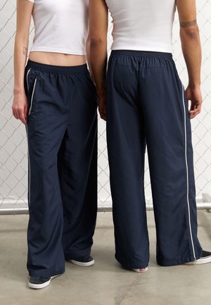 Pantalones de pierna ancha en azul marino, hechos de tejido ligero, con cintura ajustable mediante cordón, bolsillos laterales con cremallera y rayas blancas a los lados.