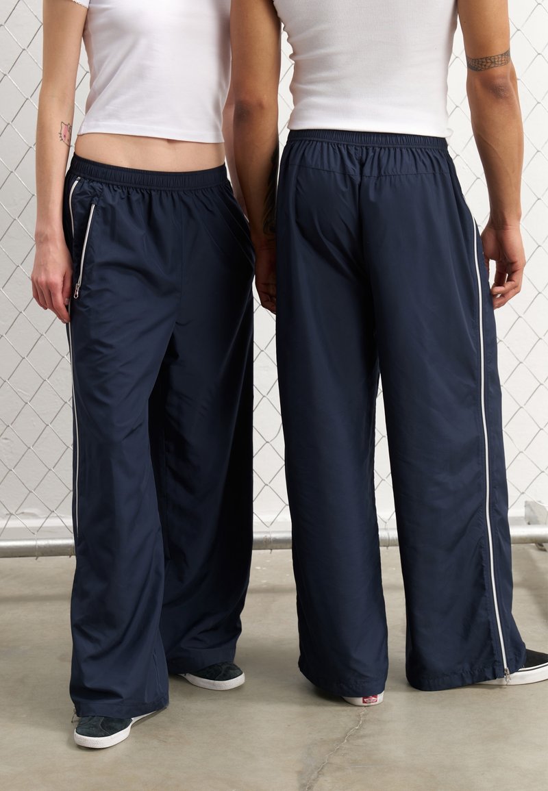 Pantalons larges bleu marine en tissu léger, avec une taille à cordon de serrage, des poches latérales zippées et des rayures blanches sur les côtés.