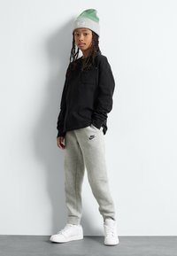 Camisa negra de manga larga, pantalones de chándal grises con el logo de Nike, zapatillas blancas y un gorro verde/gris. El modelo tiene el cabello largo trenzado.