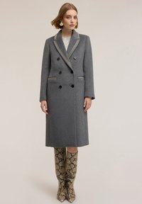 Cappotto doppiopetto grigio in lana con rifiniture a contrasto nel colletto, sei bottoni neri e una silhouette lunga. Abbinato a stivali alti fino al ginocchio con stampa a serpente.