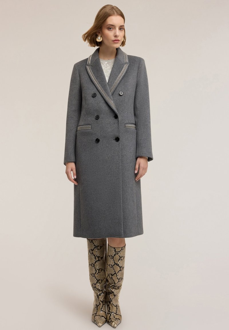 Cappotto doppiopetto grigio in lana con rifiniture a contrasto nel colletto, sei bottoni neri e una silhouette lunga. Abbinato a stivali alti fino al ginocchio con stampa a serpente.