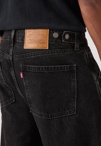 Μαύρα τζιν από ντένιμ με δερμάτινη ετικέτα που φέρει το λογότυπο της μάρκας, μεταλλικά κουμπιά και χαρακτηριστικό σχέδιο στην πίσω τσέπη.