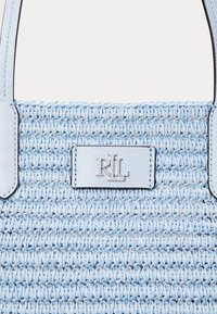 Gaiši zils austs rokas maisiņš ar teksturētu virsmu, ar tumšajiem šuvēm un metāla logo plāksni. Divi izturīgi rokturi nēsāšanai.