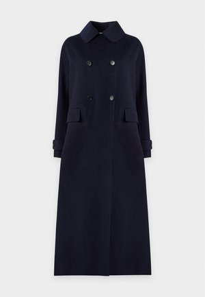 Jeune femme aux longs cheveux foncés portant un long manteau bleu marine à double boutonnage, un pull beige, un pantalon foncé et des bottines noires, debout devant un fond gris.