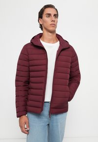 Ung mand iført en vinrød kviltet hættetrøje, hvid t-shirt og lyseblå jeans, stående med den ene hånd i lommen.