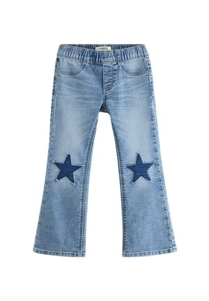 Smilende barn med krøllet hår iført en hvid t-shirt, blå jeans med stjernelapper på knæene og hvide sneakers, står legende.