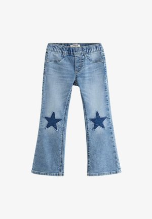 Smilende barn med krøllet hår iført en hvid t-shirt, blå jeans med stjernelapper på knæene og hvide sneakers, står legende.