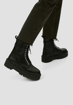 Personne portant des bottes de combat en cuir noir à lacets avec des semelles épaisses compensées et un pantalon foncé retroussé sur un fond gris clair.