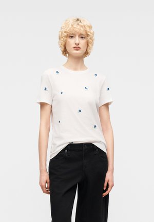 Personne blonde portant une chemise blanche à manches courtes avec de petites impressions de fleurs bleues et un pantalon noir, se tenant devant un fond uni.