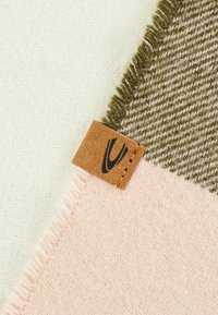 Uldblandingsstof med lyserøde, olivengrønne og cremefarvede sektioner med frynsede kanter; et lærredsmærke med logo er fastgjort.