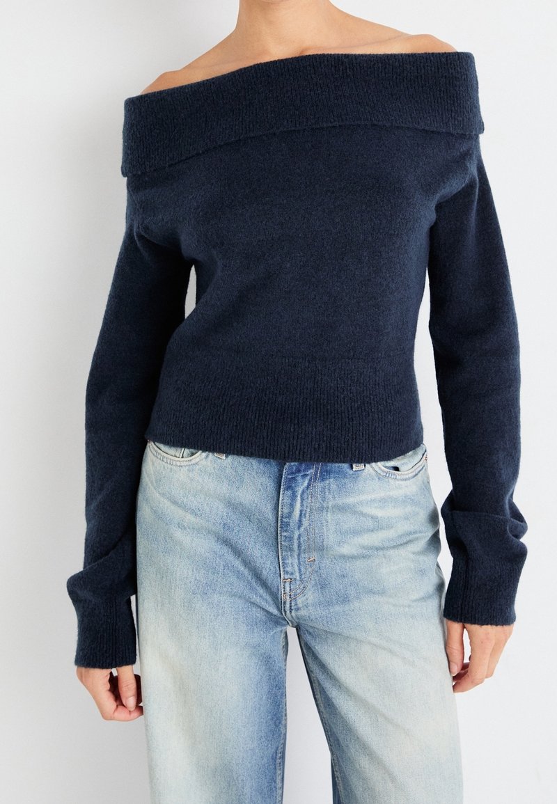 Femme portant un pull bleu marine à épaules dénudées et un jean taille haute bleu clair sur un fond uni.