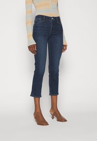 Vaqueros de mezclilla oscuros y ajustados con un dobladillo ligeramente recortado y un suave desteñido, combinados con una blusa de punto a rayas claras y mules marrones.