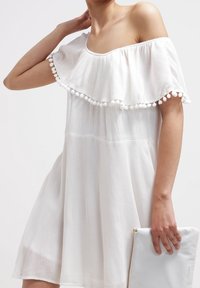 Vestido blanco sin mangas con un dobladillo de volantes en capas decorado con pompones; tejido ligero; acompañado de un bolso de mano azul claro.