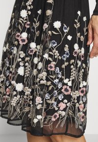 Schwarzes, transparentes Kleid aus Stoff mit gesticktem floralen Muster in Weiß, Rosa, Blau und Beige, getragen von einer Person, die eine Hand seitlich zeigt.