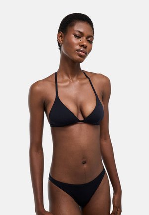 Femme aux cheveux courts portant un bikini noir, debout devant un fond clair uni, avec une expression faciale détendue.