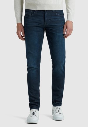 Homme portant un jean slim bleu foncé et des baskets blanches, debout devant un fond clair uni.
