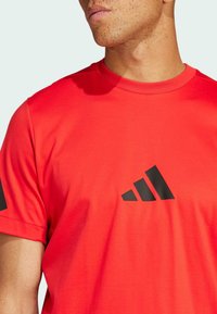 Camiseta deportiva roja hecha de tela suave con cuello redondo, que presenta un logo negro de Adidas en el pecho y detalles negros en las mangas.