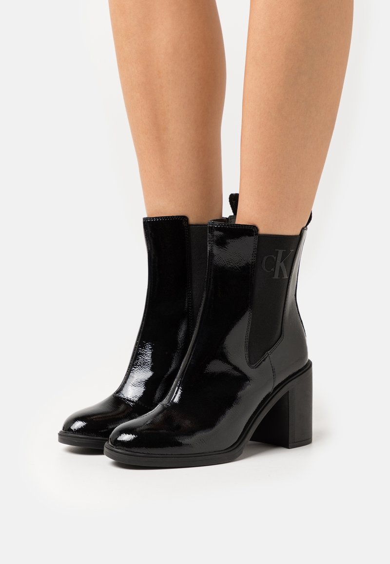 Bottines en cuir verni noir brillant à talons carrés portées sur jambes nues, avec des panneaux latéraux élastiques et un logo discret.