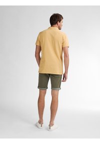 Geel polo shirt, opgerolde groene shorts, crèmekleurige sneakers. De polo heeft een klassieke kraag en korte mouwen. Eenvoudig ontwerp zonder patronen.