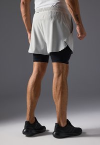 Ανοιχτό γκρι σορτς τρεξίματος με χαλαρή εφαρμογή, ελαστική μέση και πλαϊνές σχισμές, τοποθετημένα πάνω από μαύρα συμπιεστικά σορτς. Ορατά μαύρα αθλητικά παπούτσια.