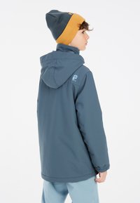 Giacca impermeabile blu con cappuccio, polsini elastici e dettagli del logo sulla spalla, abbinata a un berretto piegato blu scuro e senape.