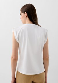Hvid ærmeløs top med struktureret skulderdesign, glat tekstur og rund udskæring. Båret med lysebrune bukser.