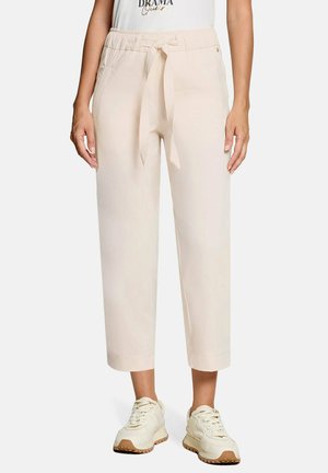 Vrouw met beige cropped broek met taillekoord, zijzakken, witte sneakers en een wit topje met gedeeltelijk zichtbare tekst.