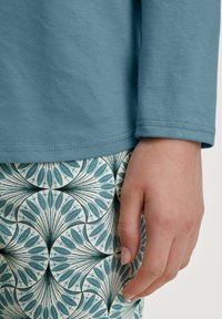 Haut à manches longues, bleu clair avec une texture lisse, associé à un tissu à motif présentant des feuilles turquoise sur un fond crème ; main visible.