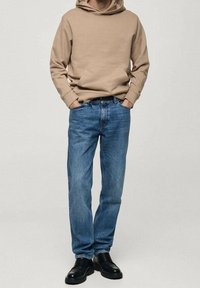 Persona che indossa un hoodie beige, jeans blu e scarpe di pelle nere, in piedi con le mani in tasca su uno sfondo chiaro e uniforme.